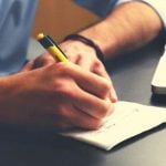 A student writing in a small notepad with a laptop next to them.