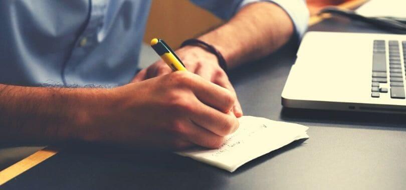 A student writing in a small notepad with a laptop next to them.