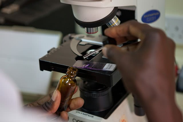 A scientist's hands were working on the microscope and holding a small container.