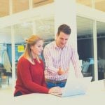 Two people standing at a desk talking while looking at a laptop.