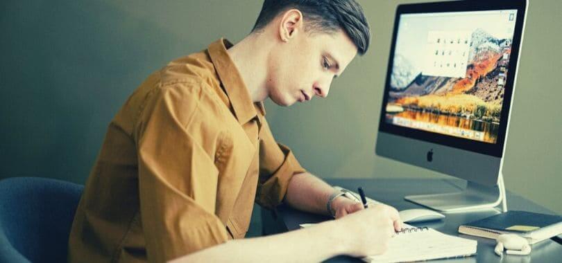 A student writing in their notebook, sitting in front of a computer.