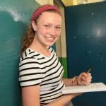 A girl smiling while holding a ballpen and notebook with both hands.