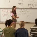 Students sitting together, looking towards a professor with a whiteboard in the background.