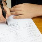 Aerial view of hand is solving a math problem on white paper with a black calculator on the desk.