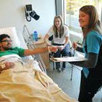 A man lying in the hospital bed, a smiling practice nurse and a woman sitting on the chair with them.