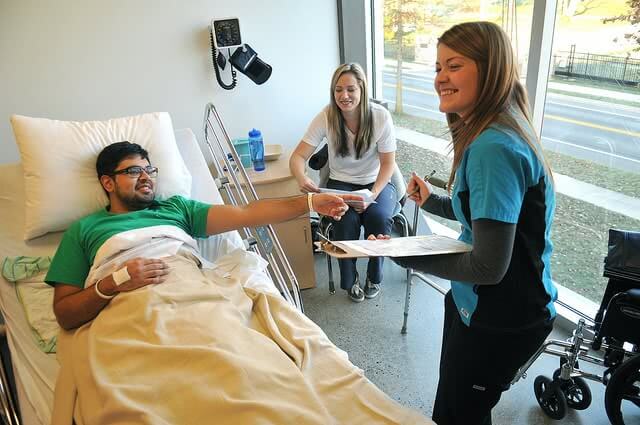 A man lying in the hospital bed, a smiling practice nurse and a woman sitting on the chair with them.