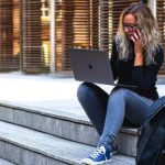 A college student sitting on steps while talking on the phone and typing on her laptop.