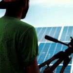 A solar photovoltaic installer holding a tool and looking out over solar panels.