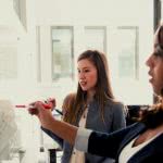 Two business administration majors writing stats on a whiteboard.