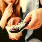 One college student eats popcorn while the other holds a remote while they watch TV.