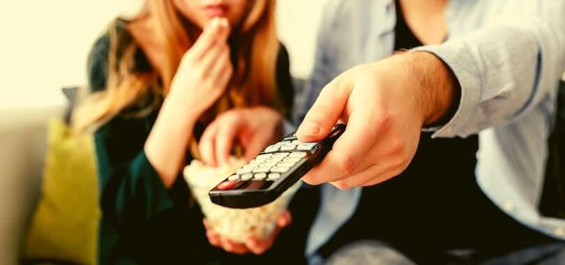 One college student eats popcorn while the other holds a remote while they watch TV.