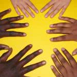 Three people of various races place their hands on a yellow table.